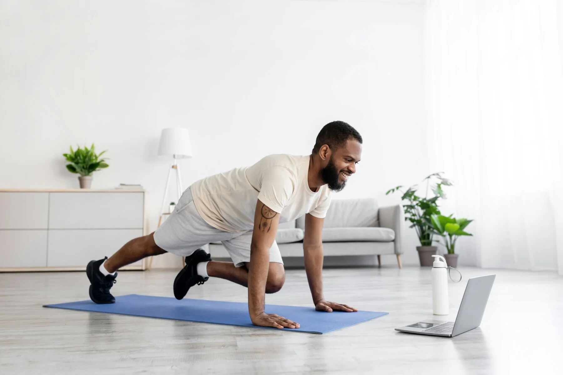 Happy young black male with beard in white sportswear do leg exercise and watch video on laptop